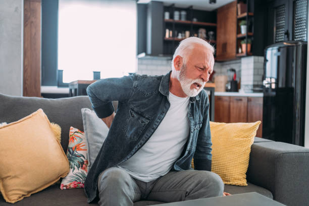 An elderly man sitting on a couch, clutching his lower back in pain.